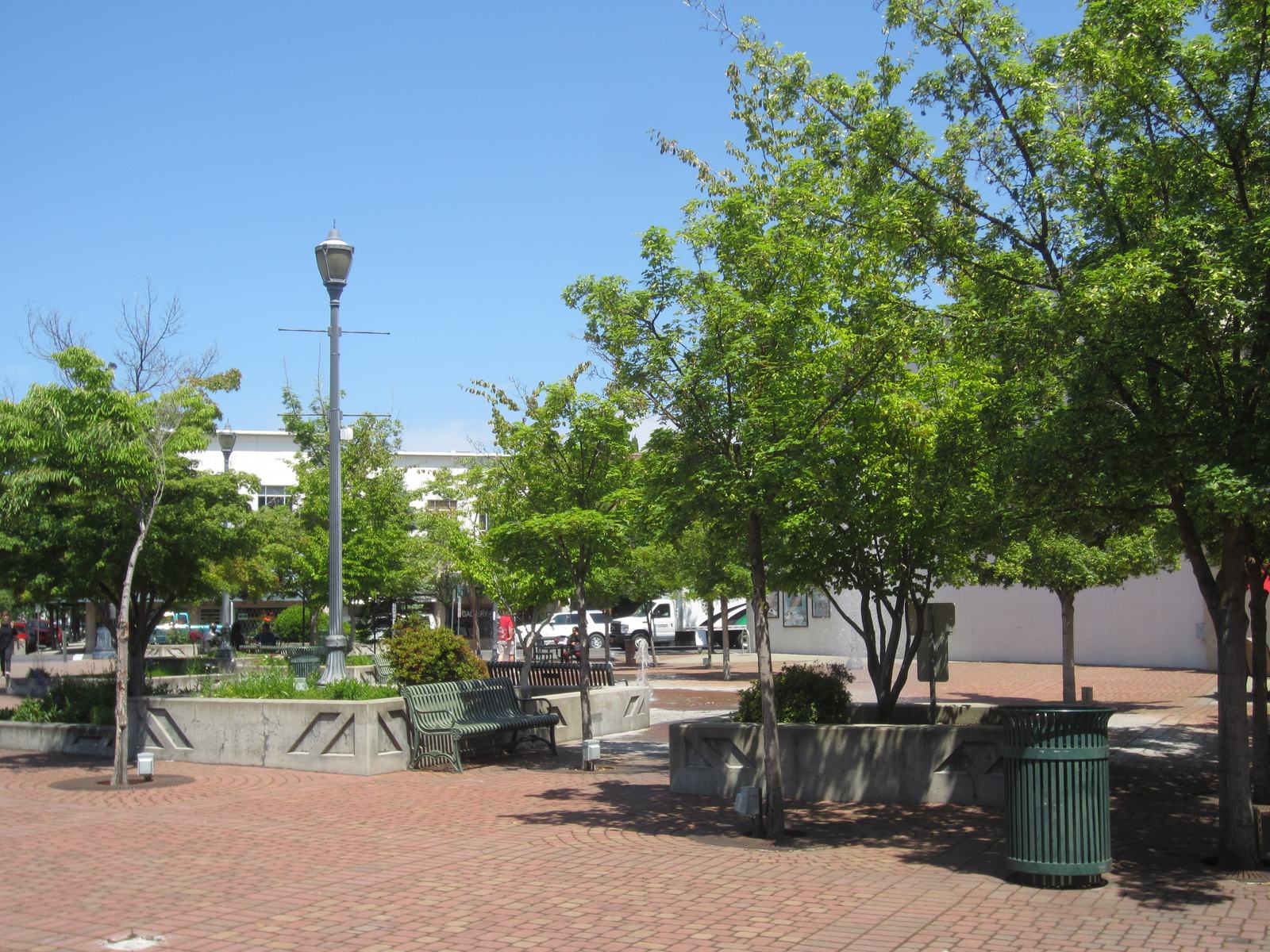 Vogel Plaza in downtown Medford, a brick public square with trees, benches, and lampposts under a blue sky